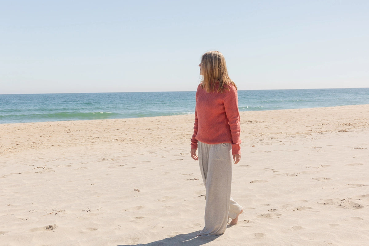  A woman stands barefoot on a sandy beach, gazing out at the calm ocean under a clear blue sky. She is wearing a cozy, coral-colored sweater from Nantucket Looms and light gray, loose-fitting pants. The scene evokes a sense of peacefulness and relaxation, showcasing the comfort and style of Nantucket Looms' apparel.